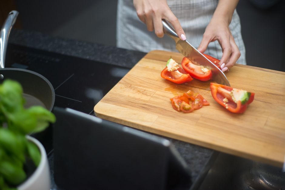 Someone slicing red peppers and tomatoes on a wooden board beside a pan and a basil plant