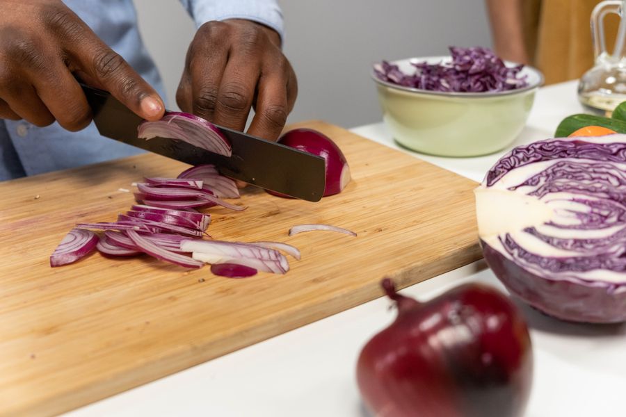 Someone slicing red onion on a wooden board beside a halved red cabbage and a bowl of shredded cabbage