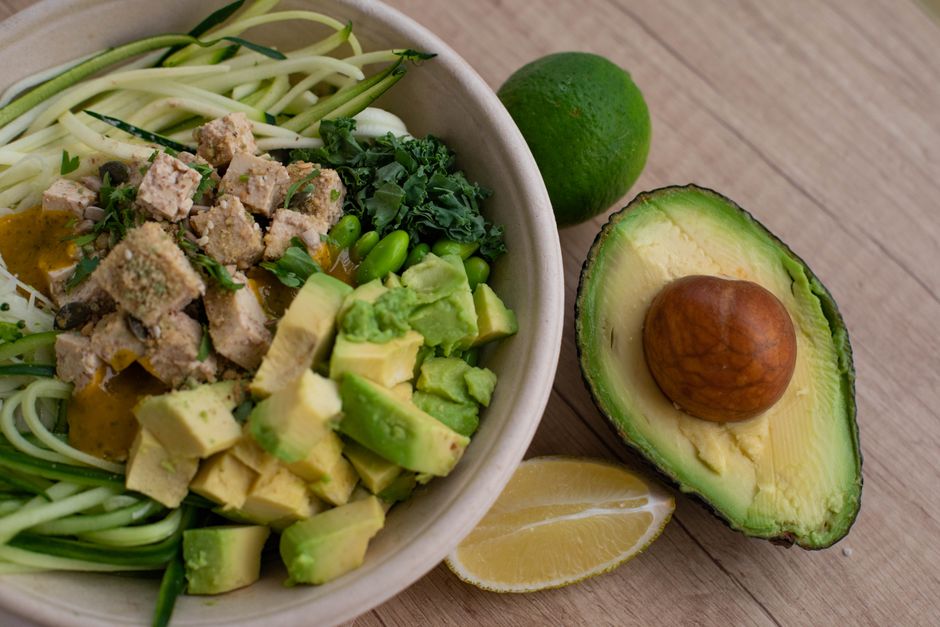 A bowl of courgette noodles with tofu, avocado, edamame and greens, beside a halved avocado and lime