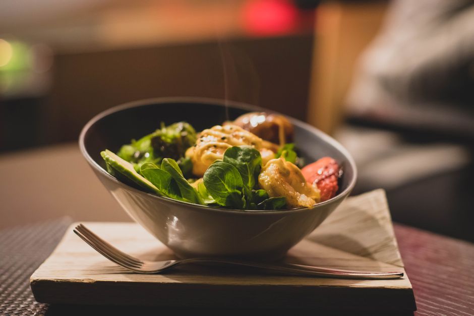 A bowl of salad leaves, avocado and roasted vegetables in a warm dressing, served with a fork on a wooden board